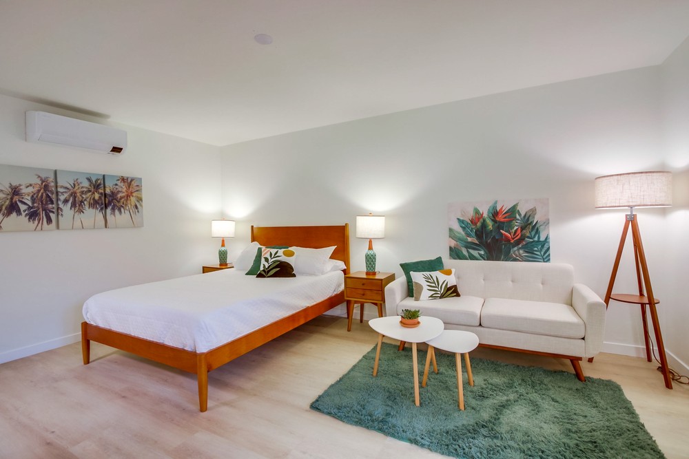 Staged studio living and sleeping area with mid-century walnut bed, sofa, palm wall art and green rug